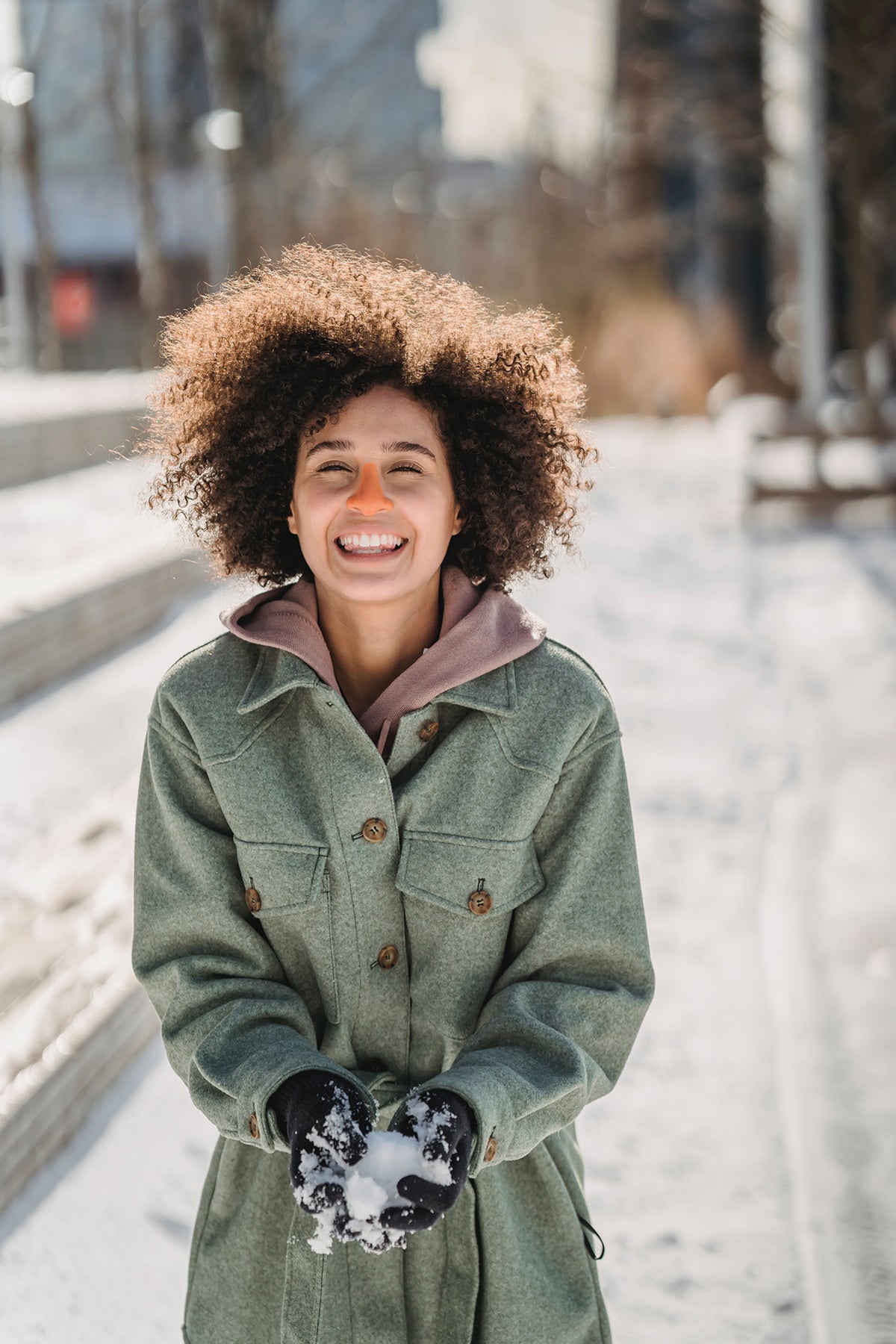 Woman wearing orange Nöz product on nose with snow in her hand in front of blurred city backround