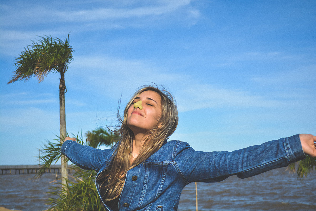 Woman wearing yellow Nöz sunscreen on nose with her arms wide and chin up, eyes closed, in front of ocean.